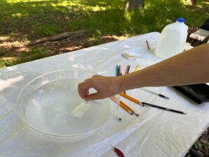 A hand places a slip of dissolving paper into a bowl of water. Paper and pens sit next to the bowl.