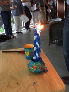 Lighted blue-and-white Havdalah candle on a wooden table, with participants' legs in background.